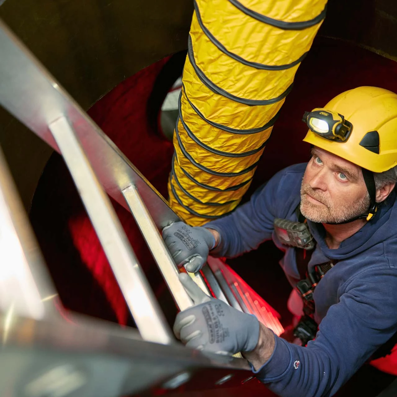 man klimmend op ladder met helm op en harnas aan in besloten ruimte