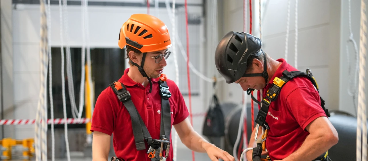 Twee personen die hun harnas goed trekken. Ze dragen een helm en staan in de trainingshal.