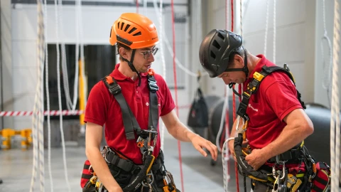 Twee personen die hun harnas goed trekken. Ze dragen een helm en staan in de trainingshal.