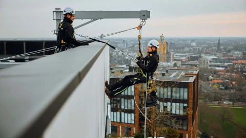 mannen aan dak hangend met touw met harnas aan en helm op